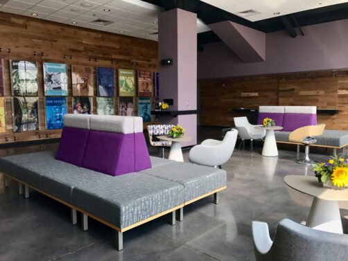 Modern lounge area at Kansas State University’s Purple Masque Theatre in Manhattan, furnished by Modern Business Interiors with bold purple seating, contemporary tables, and artistic wall displays.