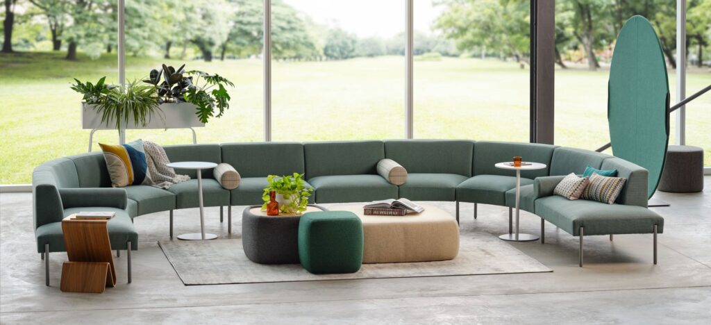 Bright workplace lounge with curved green seating, round tables, plants, and large windows