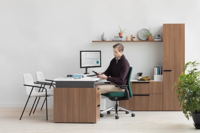 Professional seated at a modern private office desk with an ergonomic chair, computer monitor, guest seating, and wood storage cabinets.
