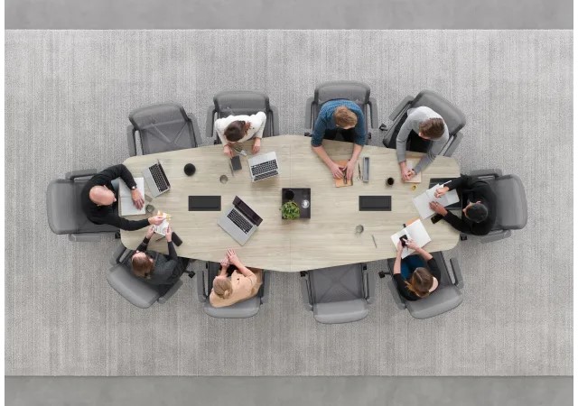 Overhead view of colleagues meeting around an oval conference table with laptops, tablets, notebooks, and office chairs.