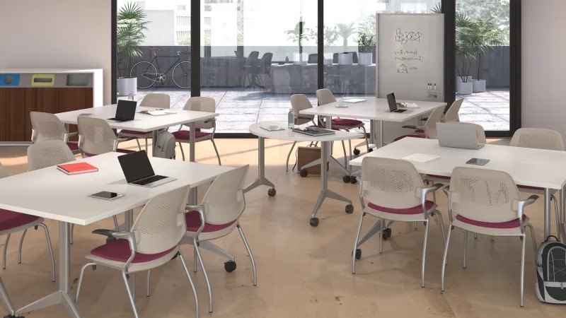 Collaborative office training space with modular tables, rolling chairs, laptops, a mobile whiteboard, and floor-to-ceiling windows.