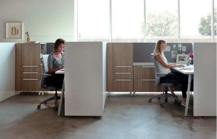 Two employees working at private office workstations with ergonomic chairs, storage cabinets, and tall dividing panels.