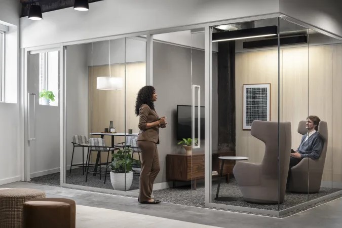 Glass-enclosed modular office with a private meeting room, high-back lounge seating, and employees collaborating.
