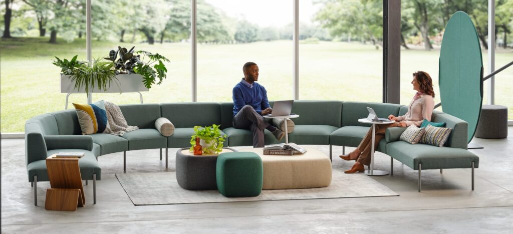 Two professionals working on laptops in a modern office lounge with modular seating, ottomans, plants, and floor-to-ceiling windows.