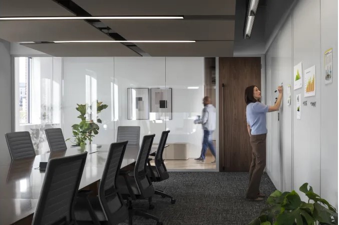 Modern conference room with ergonomic chairs, glass walls, and an employee reviewing charts on a writable wall.