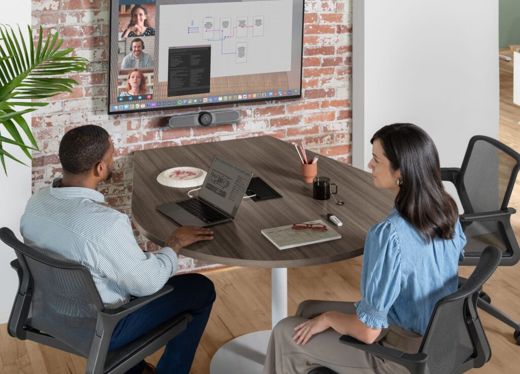 Two professionals participating in a video conference at a round meeting table with laptops and a wall-mounted display.