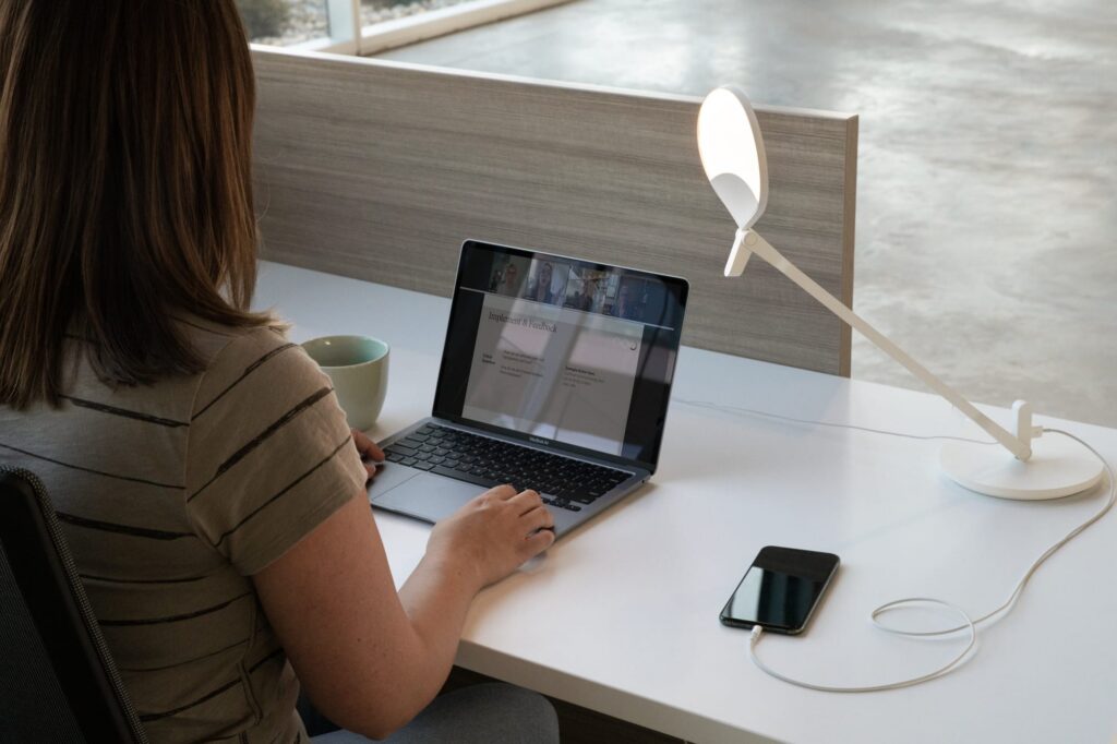 Professional attending a video meeting on a laptop at a modern workstation with a task lamp, privacy panel, coffee mug, and charging phone.