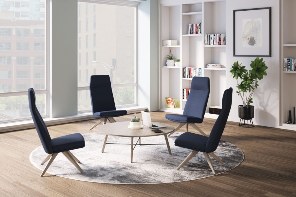 Bright office lounge with four navy high-back chairs arranged around a round coffee table, built-in bookshelves, plants, and large city-view windows.