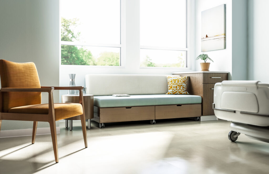 Bright healthcare patient room with a guest chair, convertible sofa, built-in storage, and hospital bed.