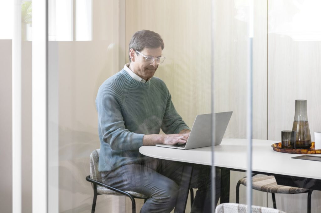 Professional working on a laptop at a table inside a bright glass-enclosed office space.