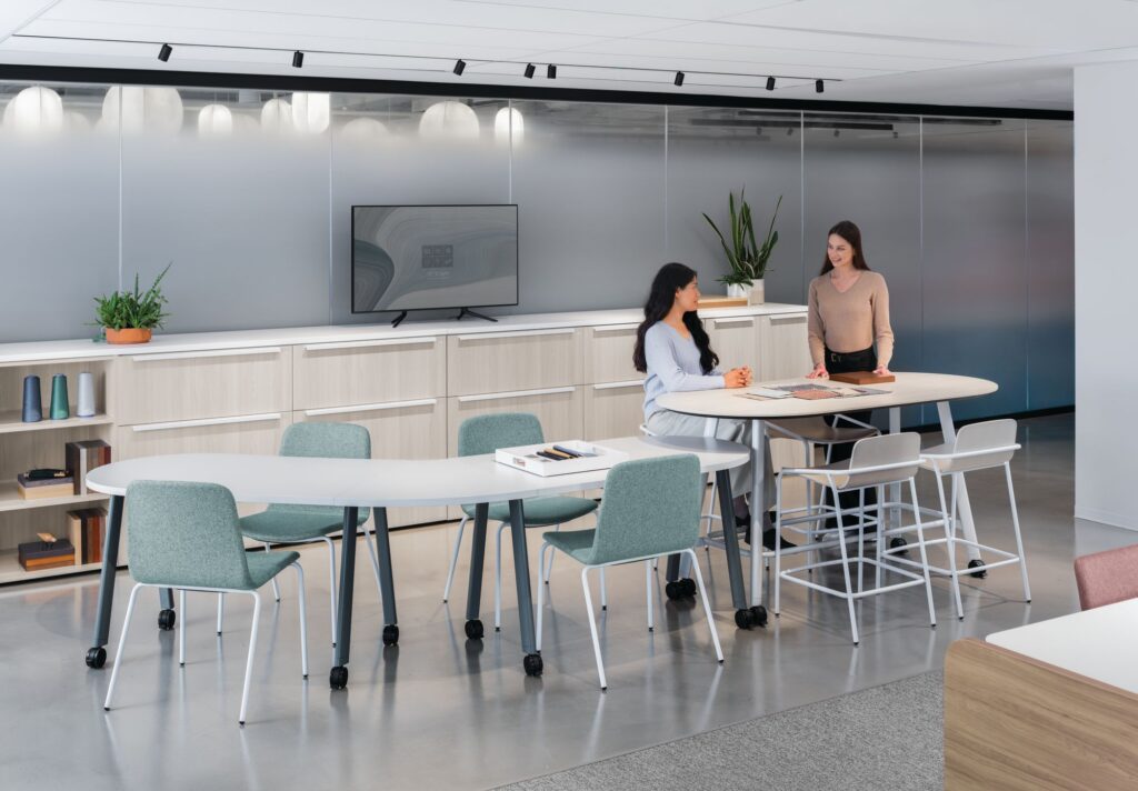 Two professionals collaborating in a flexible office workspace with mobile tables, mixed-height seating, built-in storage, and a wall-mounted display.