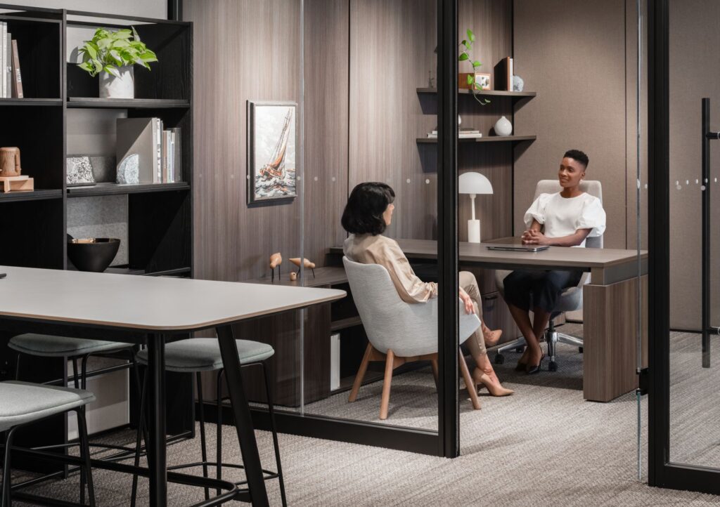 Two professionals meeting in a glass-enclosed private office with wood furnishings, upholstered chairs, built-in shelving, and an adjacent collaboration table.