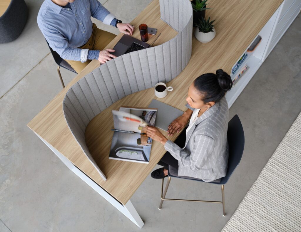 Overhead view of two professionals working at a shared wood table with curved acoustic privacy dividers, laptops, plants, and office materials.
