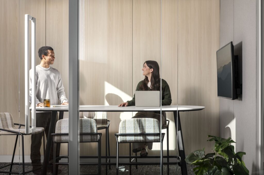 Two professionals collaborating at a high table with a laptop inside a glass-enclosed meeting room.