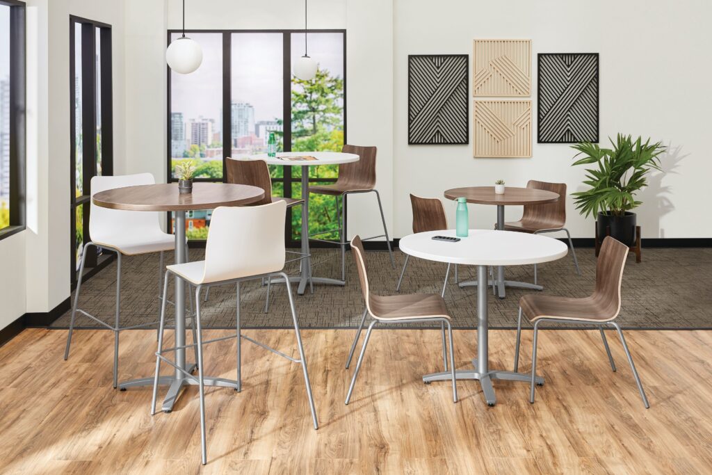 Modern office café with round tables, mixed-height seating, pendant lights, wall art, and large windows overlooking the city.