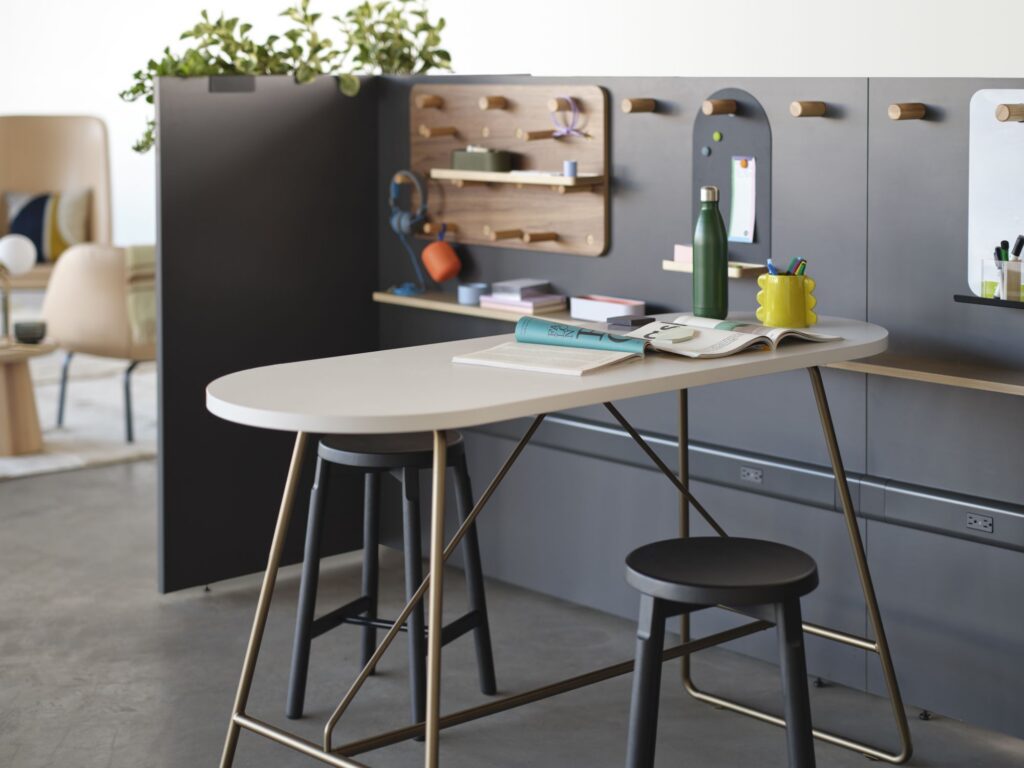 Modern collaborative workspace with a counter-height table, stools, integrated wall storage, pegboard accessories, plants, and office supplies.