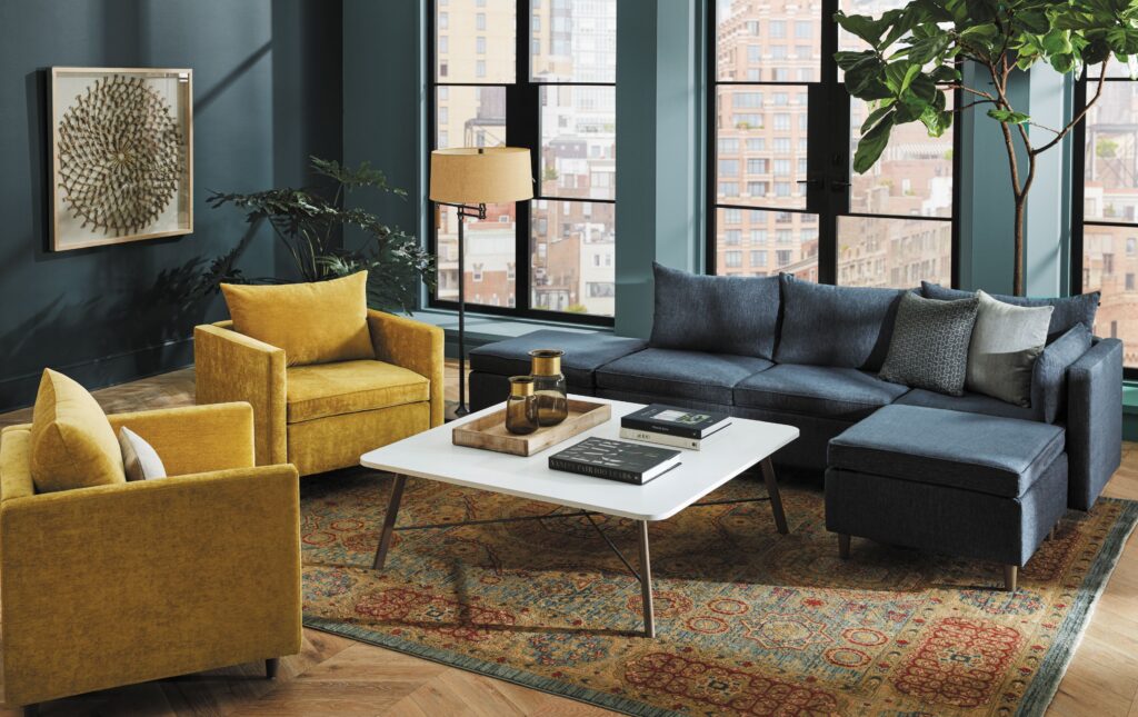Stylish office lounge with a navy sectional sofa, mustard armchairs, coffee table, patterned rug, indoor plants, and city-view windows.