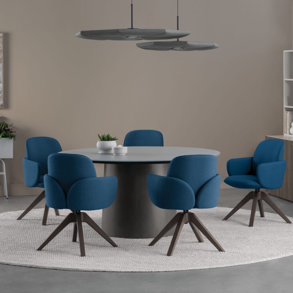 Modern meeting area with a round conference table, blue upholstered swivel chairs, suspended acoustic lights, and a neutral office interior.
