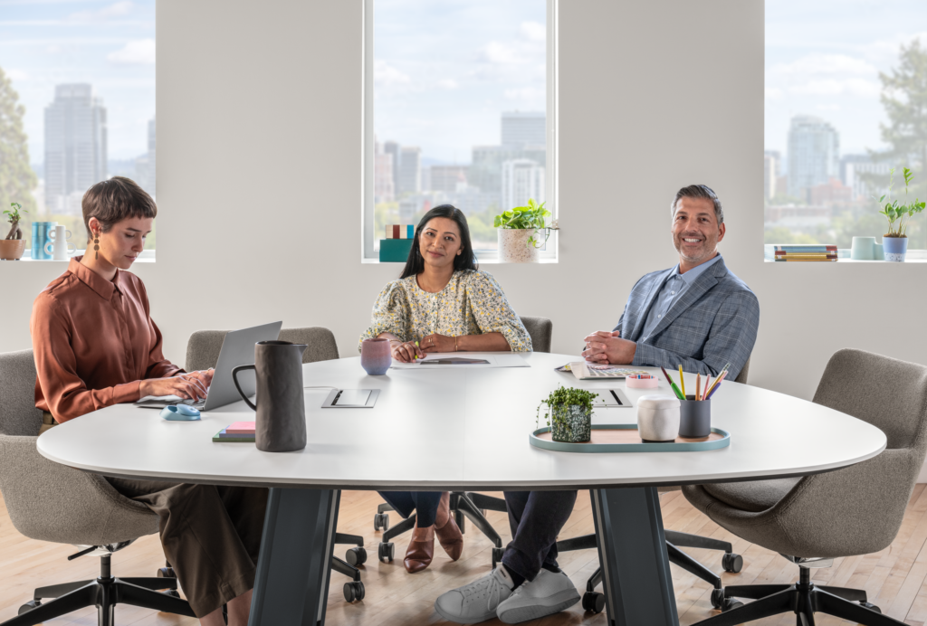 Three professionals meeting around a large conference table with a laptop, office supplies, upholstered chairs, and city-view windows.