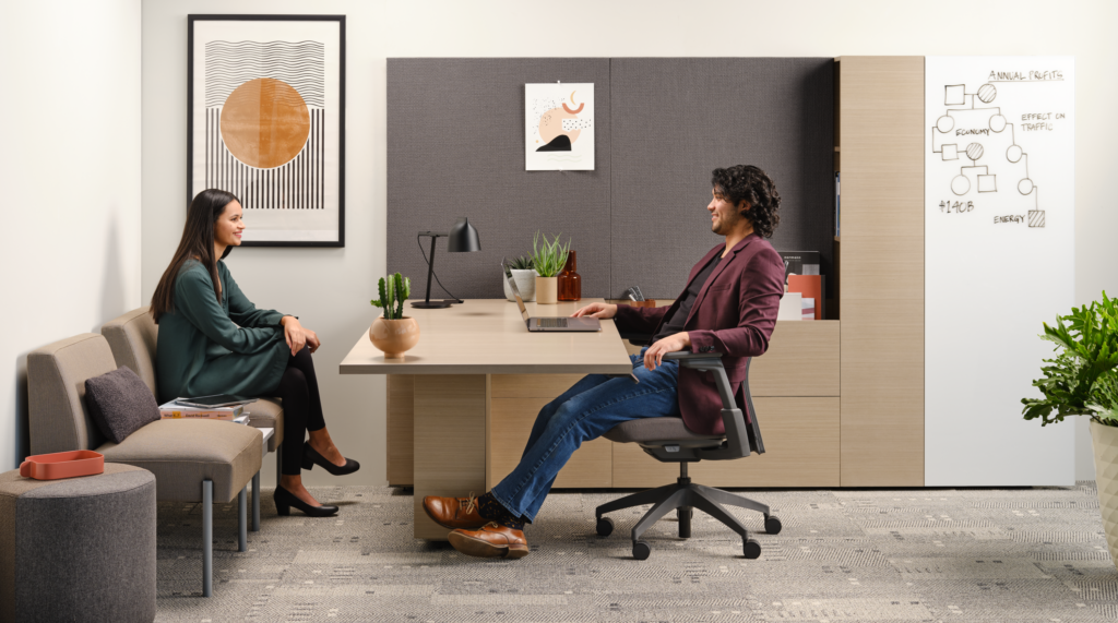 Two professionals meeting in a private office with a wood desk, ergonomic seating, lounge bench, acoustic wall panels, and a writable planning wall.