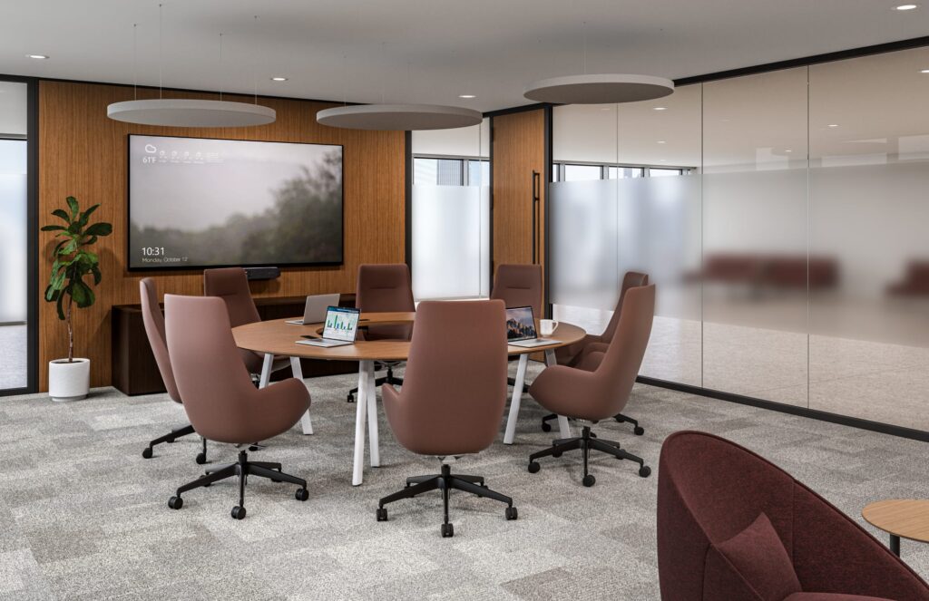Executive conference room with a round wood meeting table, upholstered swivel chairs, wall-mounted display, glass partitions, and suspended acoustic panels.