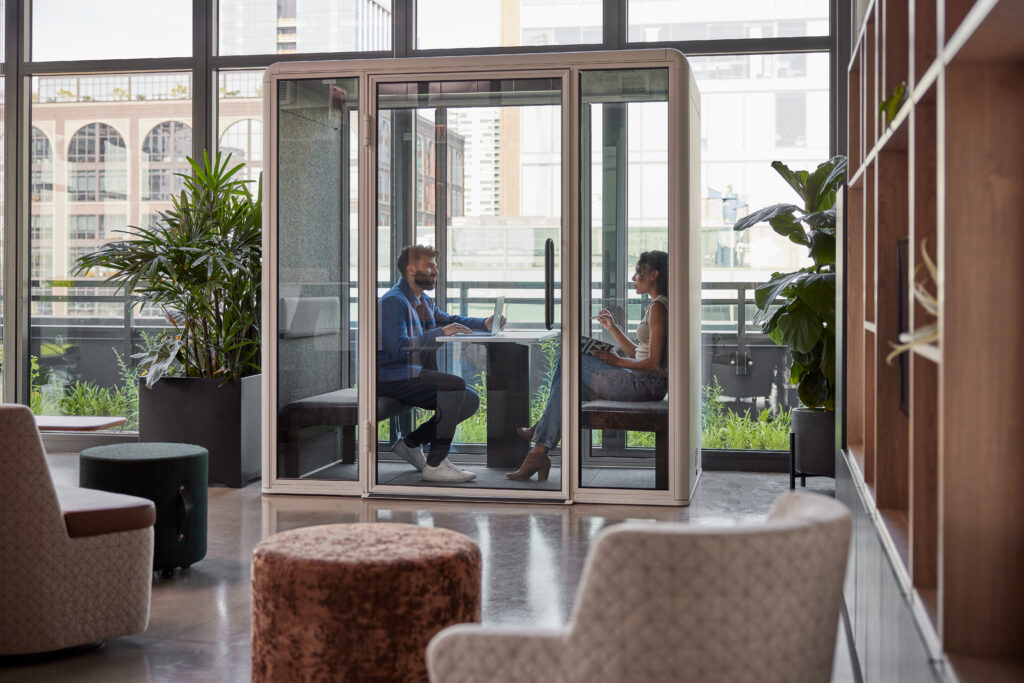 Coworkers having a private discussion inside a freestanding glass meeting pod in a modern office lounge.